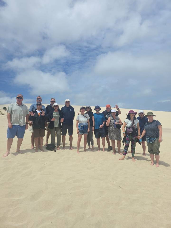 Tour group stands on wide sand dunes under partly cloudy sky, some waving at camera.