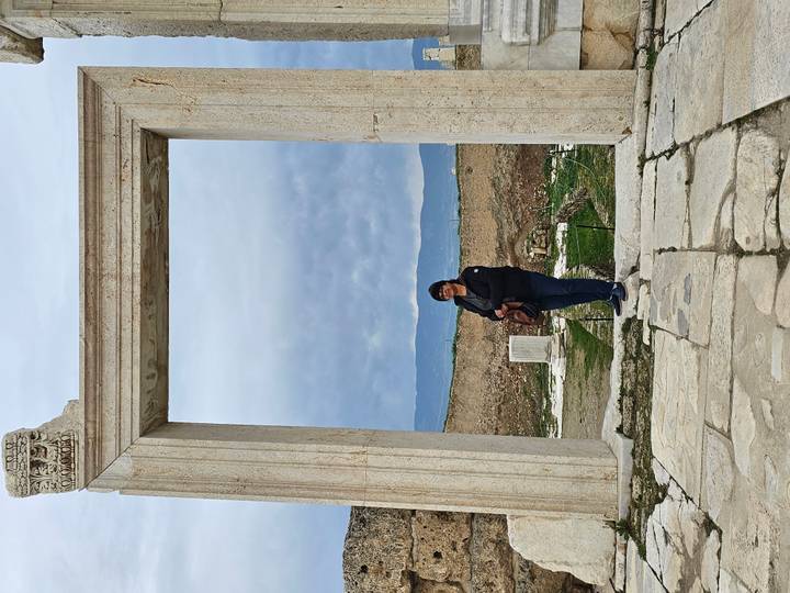 Tourist stands framed by a tall marble archway among ancient ruins with mountains in the distance.