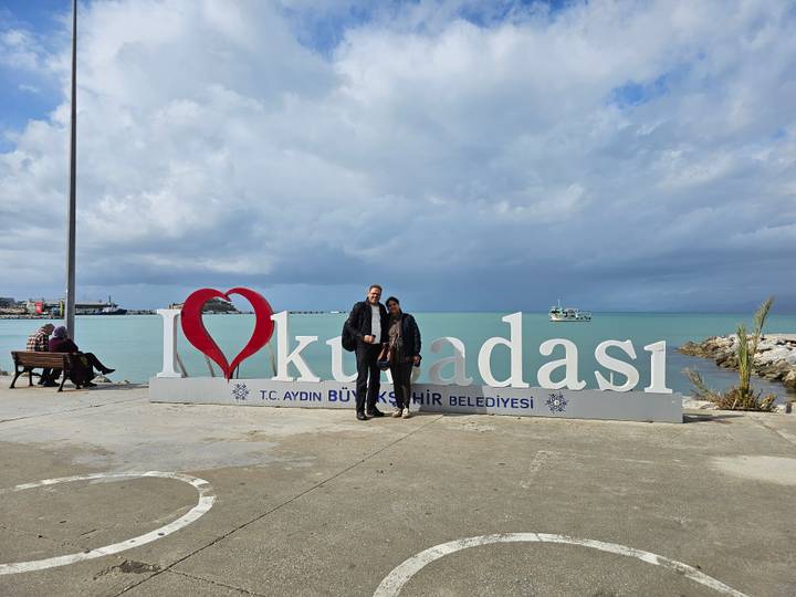Couple poses before large seaside sign reading 'I ❤️ Kusadasi' with turquoise water and ferry behind.