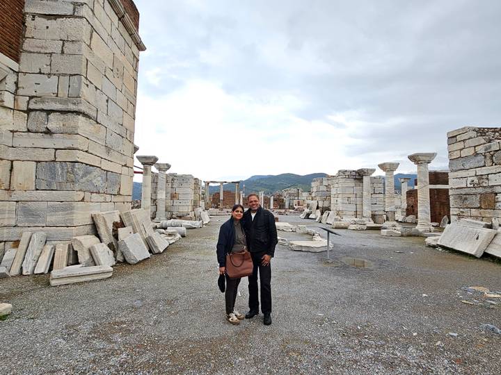 Two travellers stand amid scattered marble columns and ruins of an early Christian basilica.