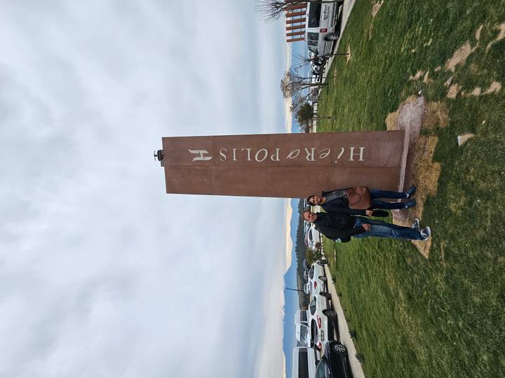 Visitors stand beside a tall brown stone monument marked 'Hierapolis' on a grassy plain.