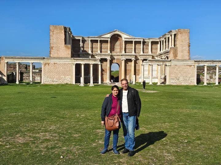 Couple stands on grassy forecourt before the grand colonnaded façade of the ancient Sardis gymnasium.