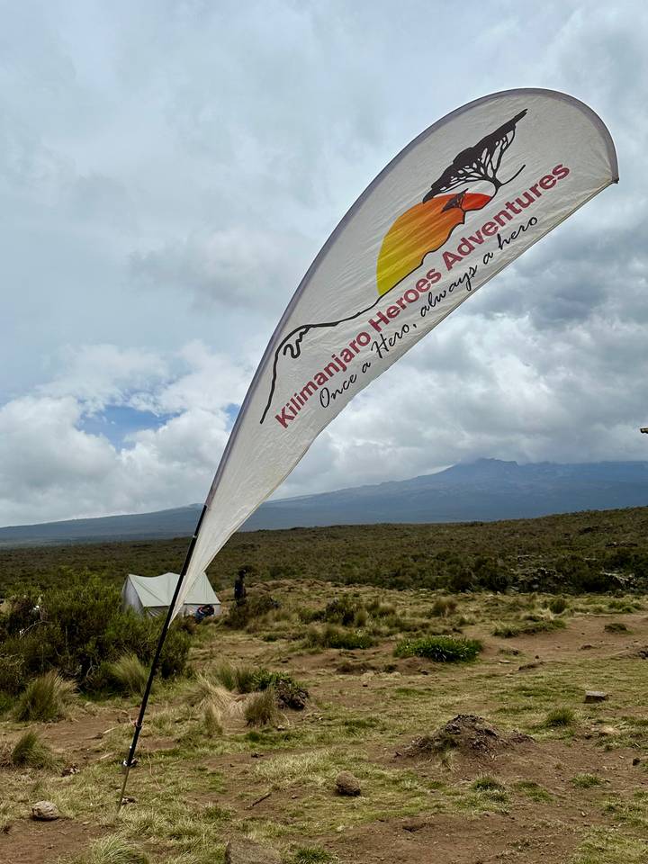 Flag bearing Kilimanjaro Heroes logo fluttering above a campsite with Mount Kilimanjaro in the distance