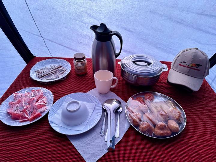Camping meal setup inside a tent with covered pastries, fruit, thermos and utensils on a red cloth