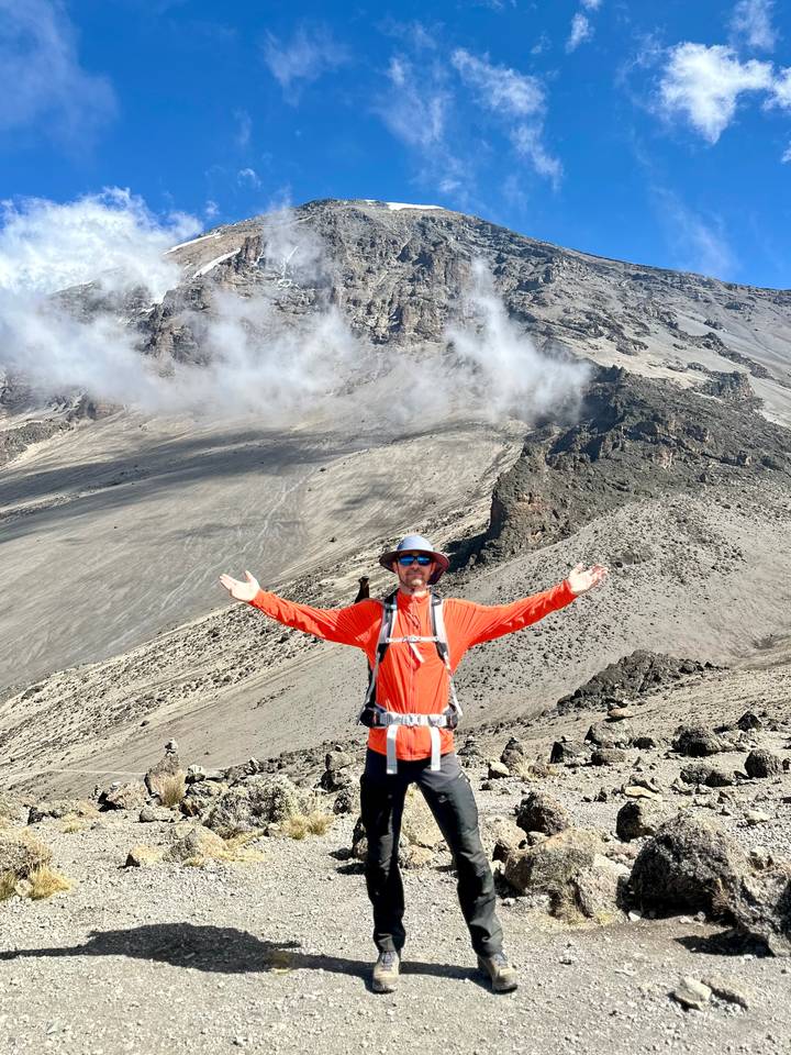 Trekkers stands with arms outstretched on a volcanic slope of Mount Kilimanjaro with clouds swirling below
