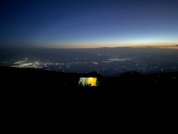 Glowing tent on a mountainside overlooking a city’s lights under a deep blue sunset sky.