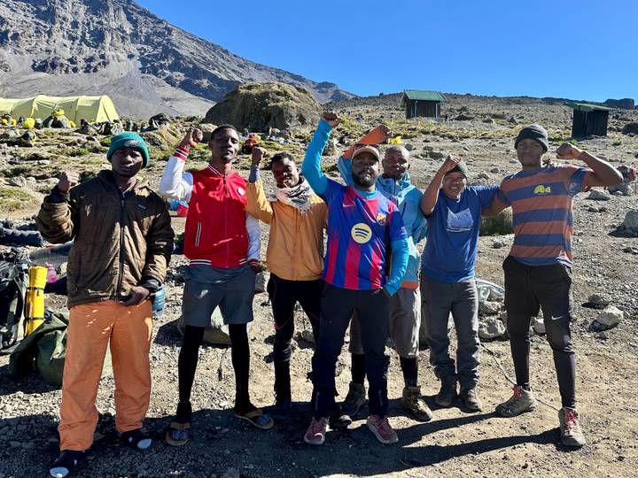 Seven cheerful mountain guides pose with raised fists at a high-altitude campsite.