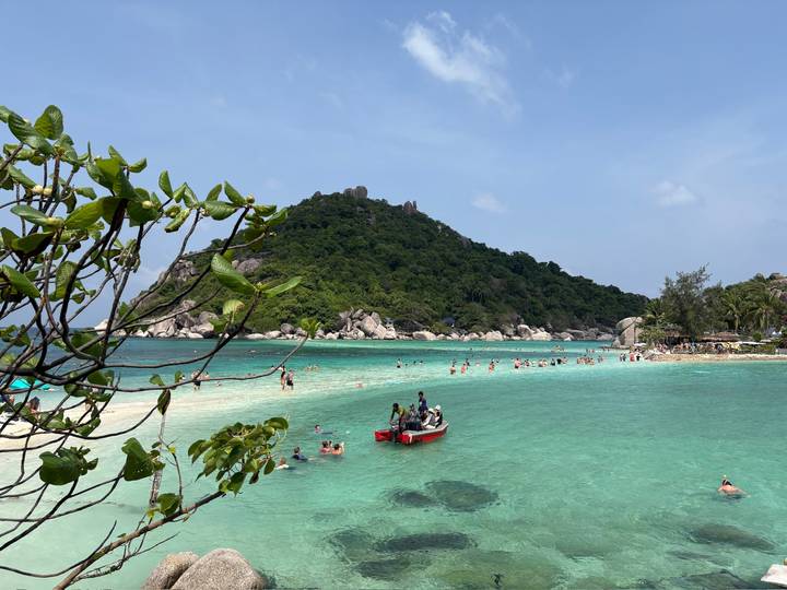 Turquoise bay with swimmers and a small red boat, framed by a lush green island under blue sky.