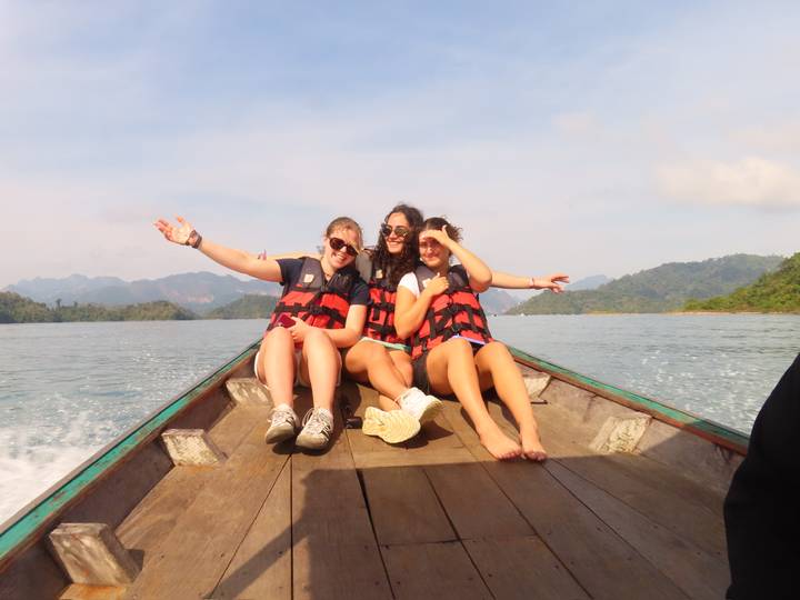 Three young women in life vests relax on the bow of a wooden long-tail boat cruising a lake surrounded by mountains.