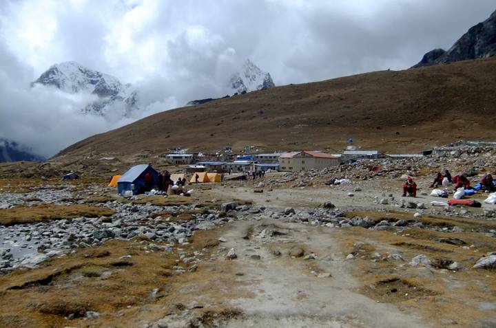 Mountain hamlet with trekkers, tents and stone lodges beneath partially cloud-covered Himalayan peaks.