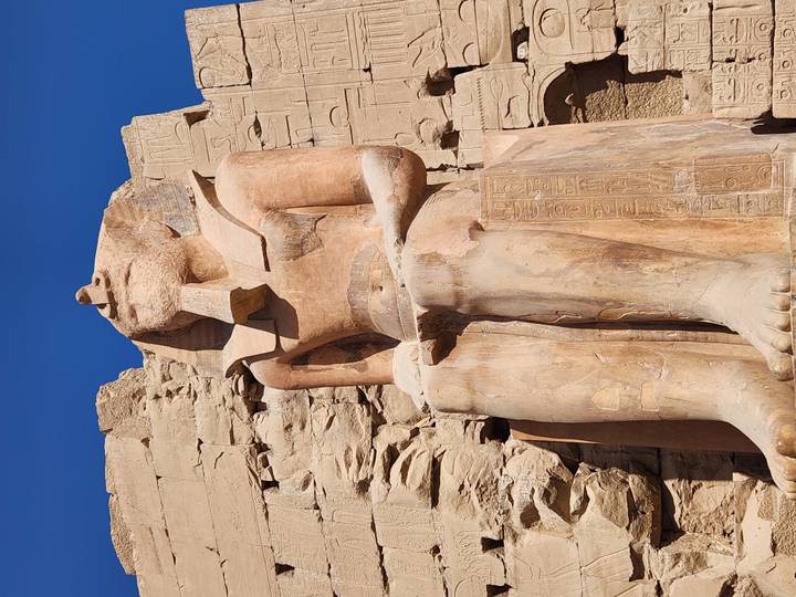 Upward view of a seated pharaoh statue carved in sandstone against a vivid blue sky.