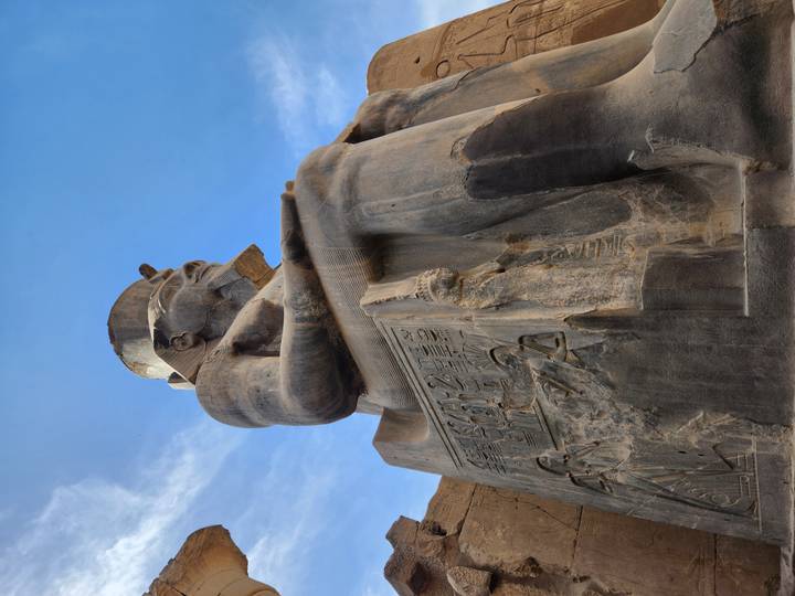Low-angle view of a colossal granite statue of a seated pharaoh against a bright sky.