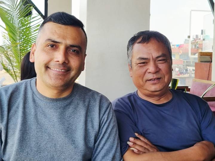 Two men smiling for the camera inside a casual café with cityscape glimpsed through the window.