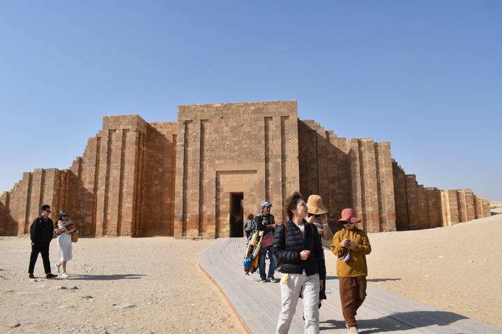 Tourists walk toward the stepped entrance building at Saqqara on a sandy desert floor.