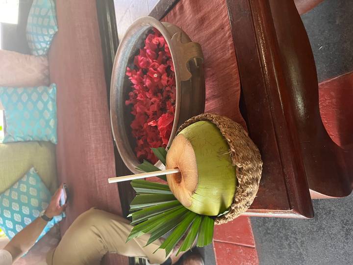 Fresh coconut drink served on a woven coaster beside a bowl of rose petals on a wooden table.
