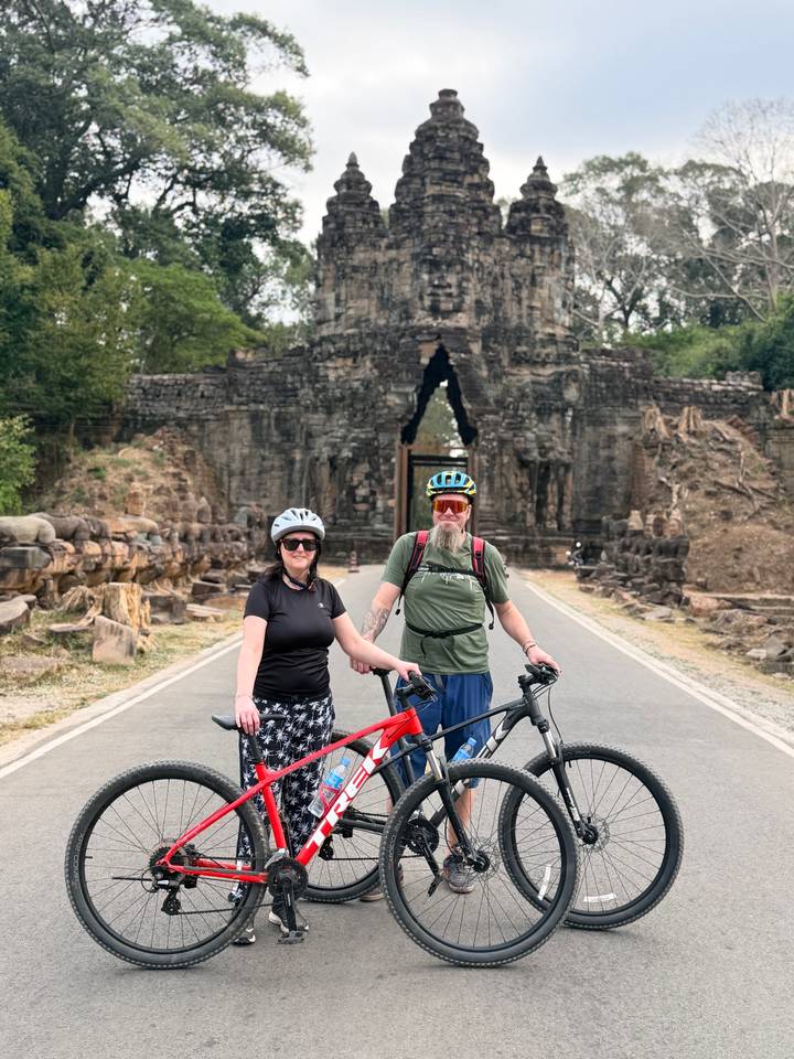 Cyclists pose with bikes on road leading to ancient Angkor stone gate lined with statues.