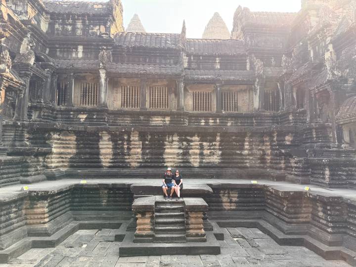 Couple sits on ancient stone steps inside Angkor Wat cloister surrounded by weathered carvings.