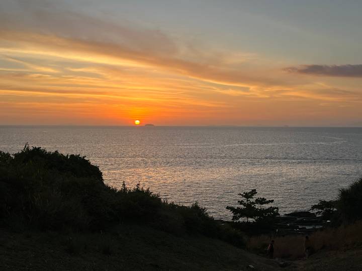 Orange sun sets over a calm sea viewed from a coastal hillside.