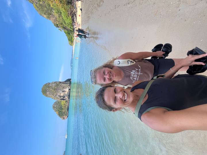 Two women smile for a selfie on Railay Beach with clear water and a tall karst cliff behind.