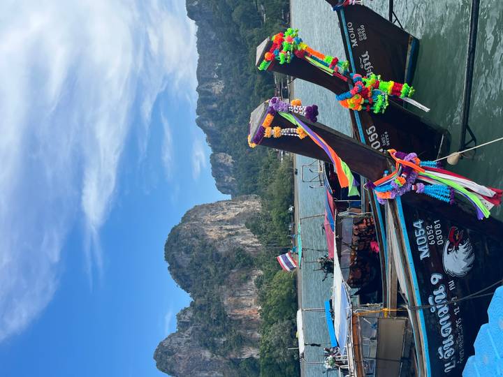 Colourful decorated long-tail boats float on green water with towering limestone cliffs in the background.
