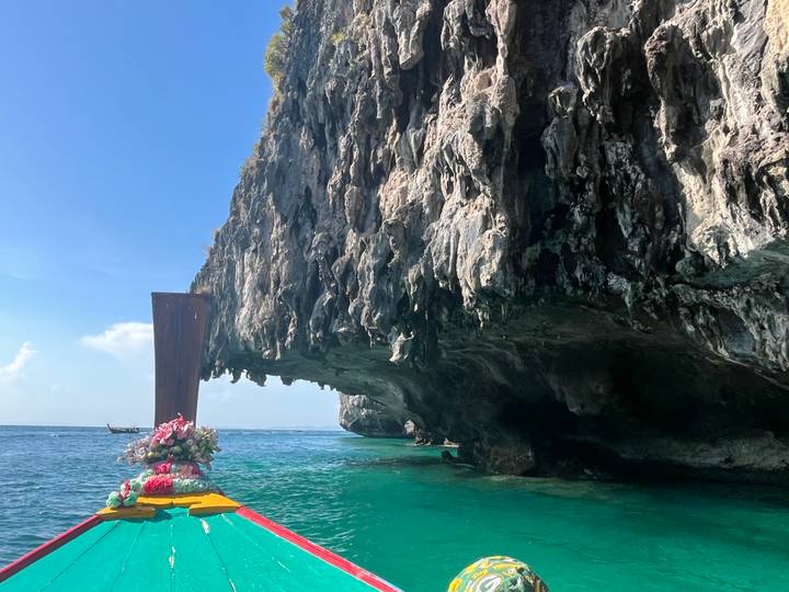View from the bow of a long-tail boat approaching a dramatic overhanging limestone cliff over turquoise water.