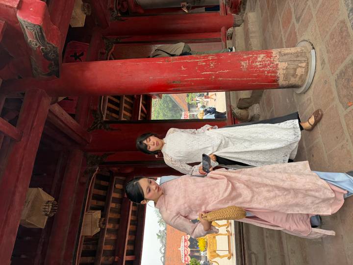 Two women in elegant traditional Ao Dai walk through red-painted temple pillars.