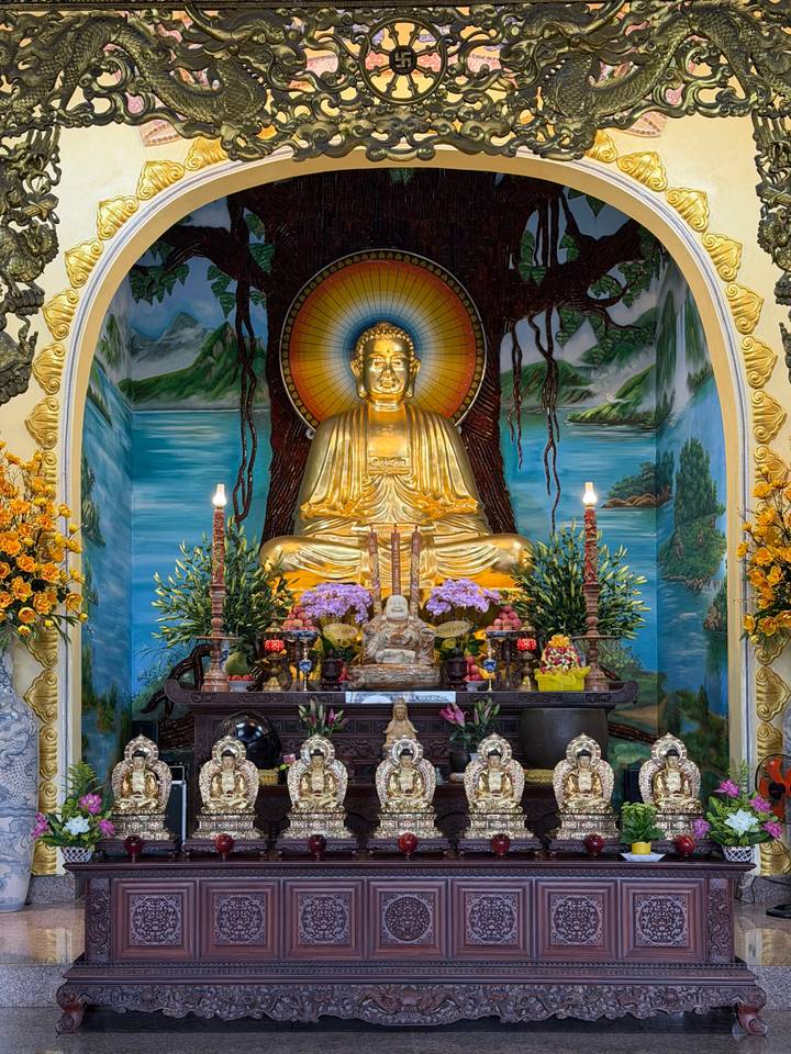 Vibrant altar with golden seated Buddha surrounded by offerings, flowers and murals.