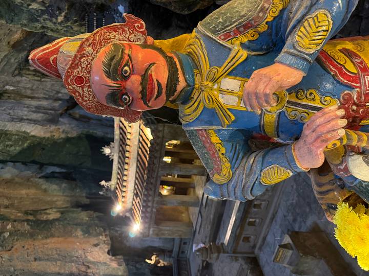 Colorful guardian statue inside a dimly lit cave temple with stone roof in background.