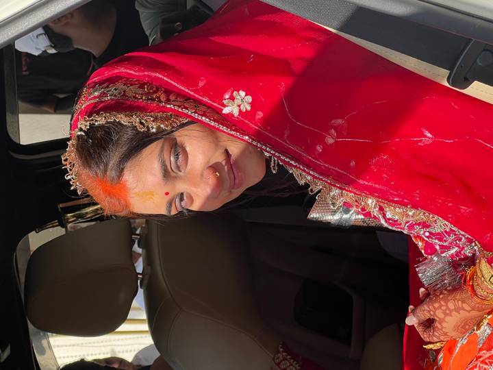 Close-up of a bride in a red sari and jeweled headpiece inside a vehicle, lit by strong sunlight.