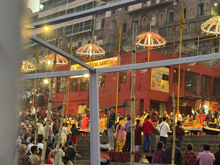 Evening Ganga Aarti ceremony with priests and large crowds outside a red riverside shrine lit by umbrellas of lights.