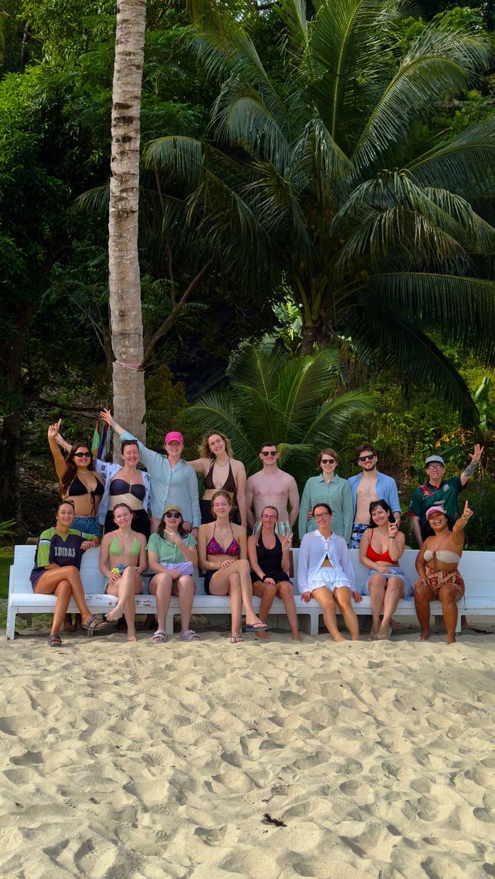A large group of swimsuit-clad travellers posing together on a bench framed by tropical palm trees and jungle greenery.