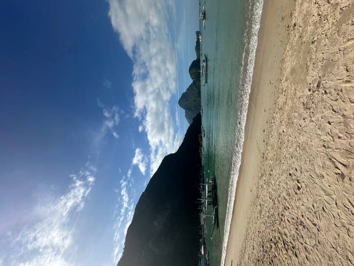 A tranquil tropical beach with soft sand, emerald water, fishing boats and dramatic limestone mountains under a clear blue sky.