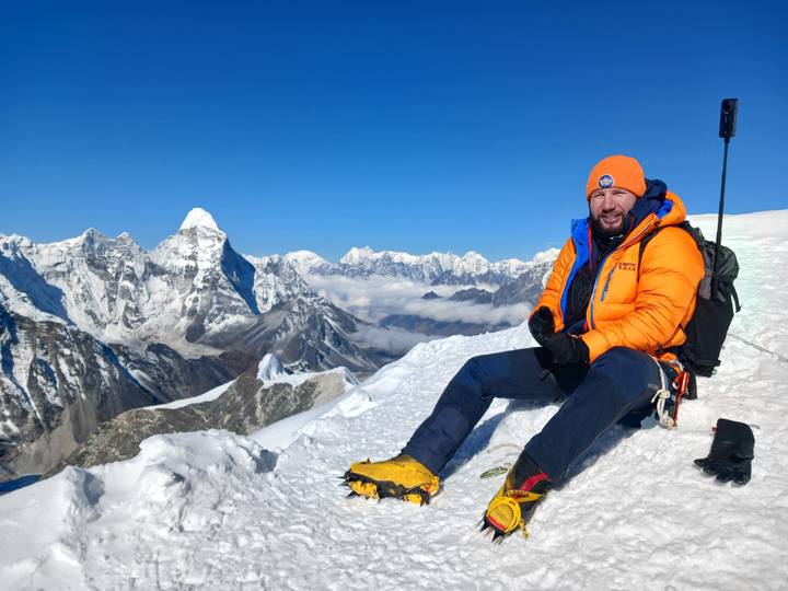 Mountaineer in bright orange jacket and crampons rests on a snowy summit ridge with vast Himalayan panorama.