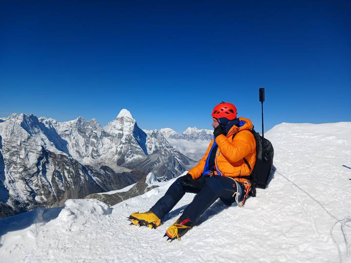 Climber wearing a red helmet sits on a snowy peak, admiring the formidable Himalaya under a clear sky.