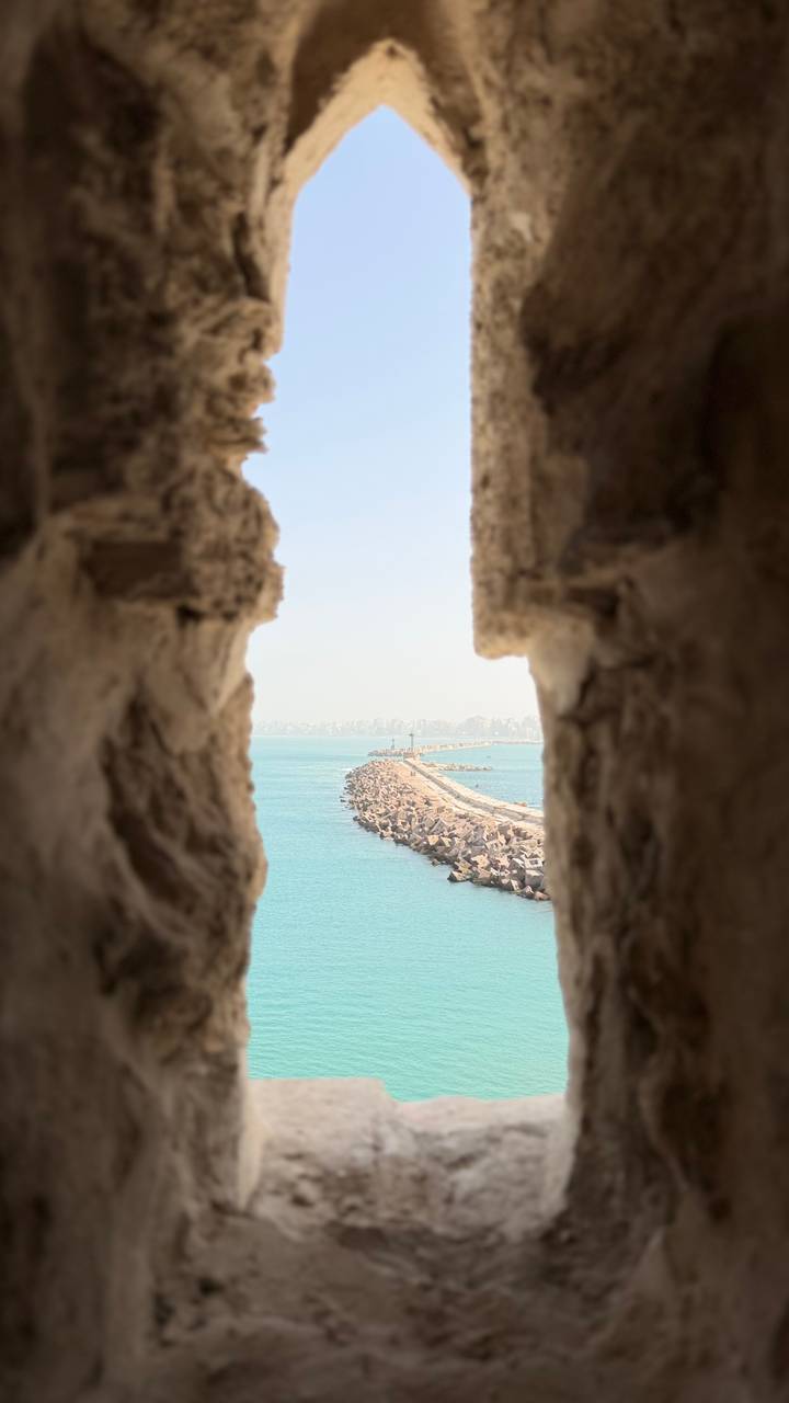 View of a long stone breakwater and distant city skyline framed by weathered castle walls over turquoise water.