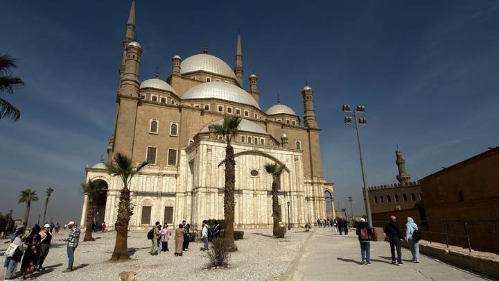 Grand Mosque of Muhammad Ali with soaring minarets and domes stands against a blue Cairo sky as tourists explore the forecourt.