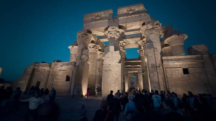 Nighttime visit to Kom Ombo Temple where spotlights illuminate the massive columns against a deep blue sky as visitors walk below.