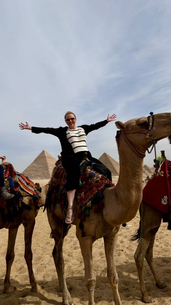 Tourist joyfully spreads arms while seated on a camel with pyramids behind