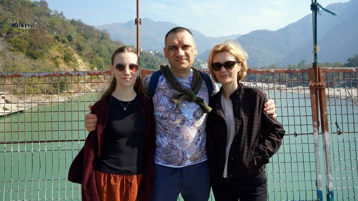 Three travelers pose on a suspension bridge over a turquoise river with mountains beyond