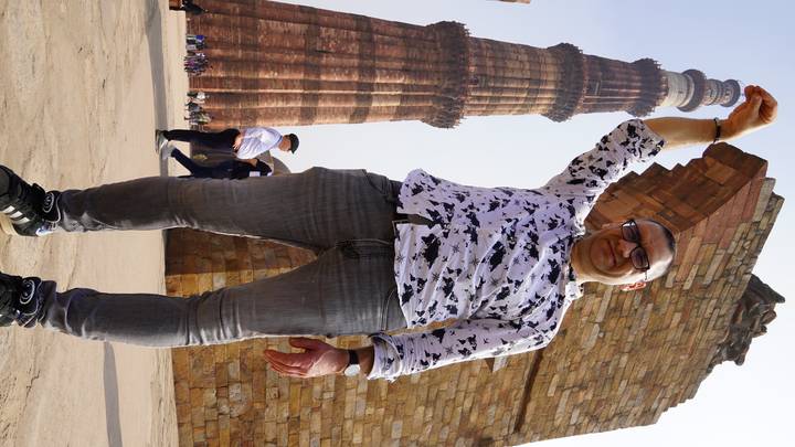 Man playfully poses as if holding the towering Qutub Minar while others walk nearby