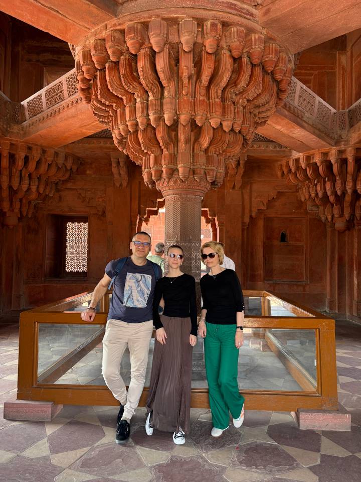 Travelers pose inside an ornately carved red sandstone hall with intricate ceiling