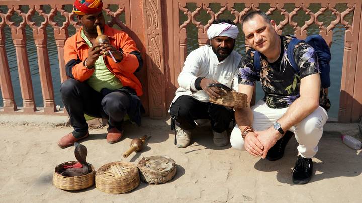 Two snake charmers sit on a pavement with baskets and a cobra while a tourist crouches beside them.