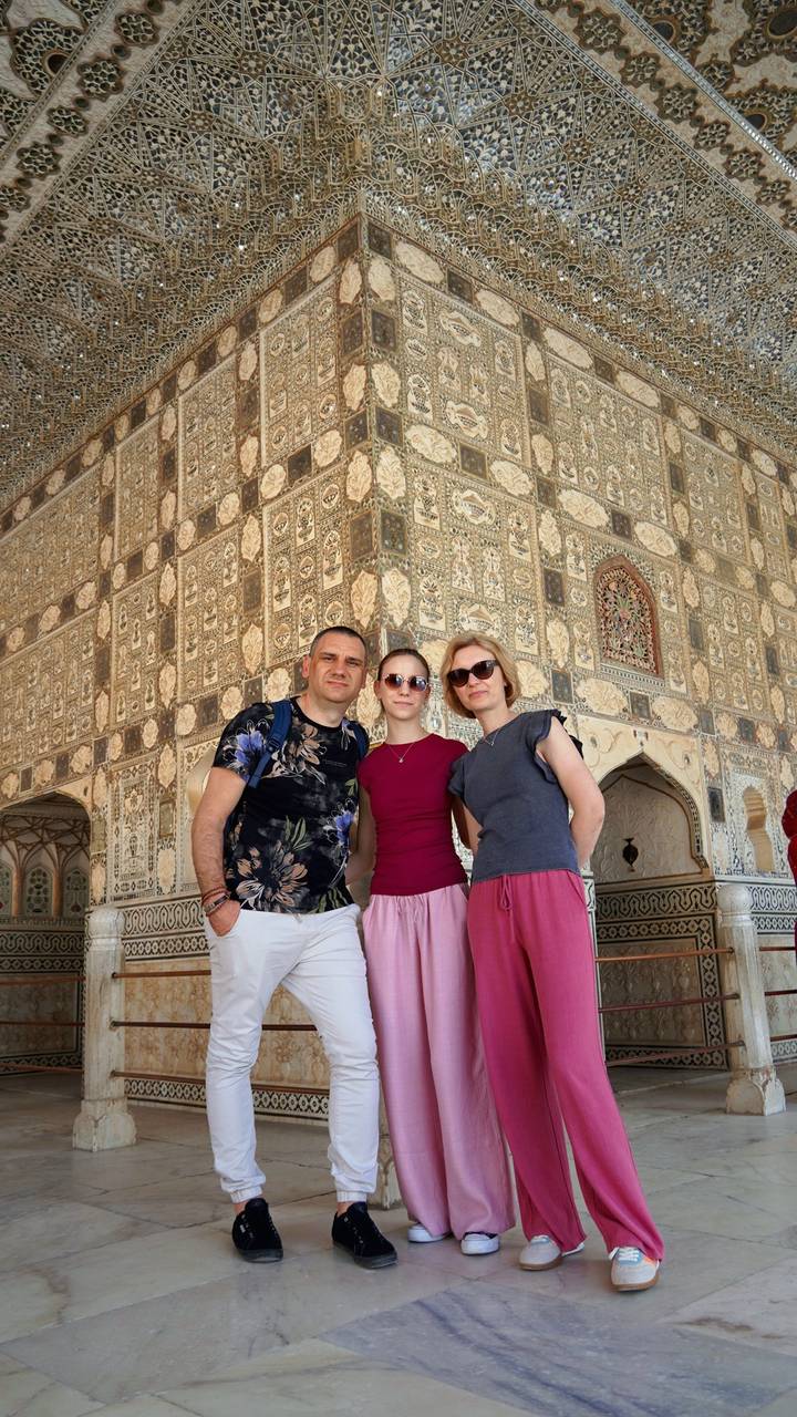 Three travellers smile in front of a richly decorated mirror-work wall inside Amber Fort.