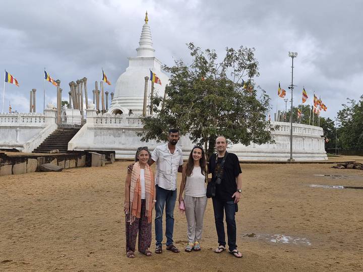 Four travellers pose in front of a large white stupa with prayer flags and ancient columns.