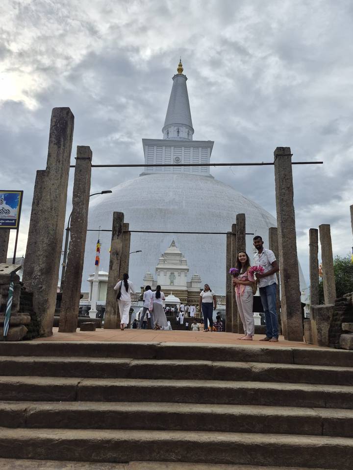 Pilgrims and a smiling couple hold lotus flowers before a giant stupa surrounded by pillars.