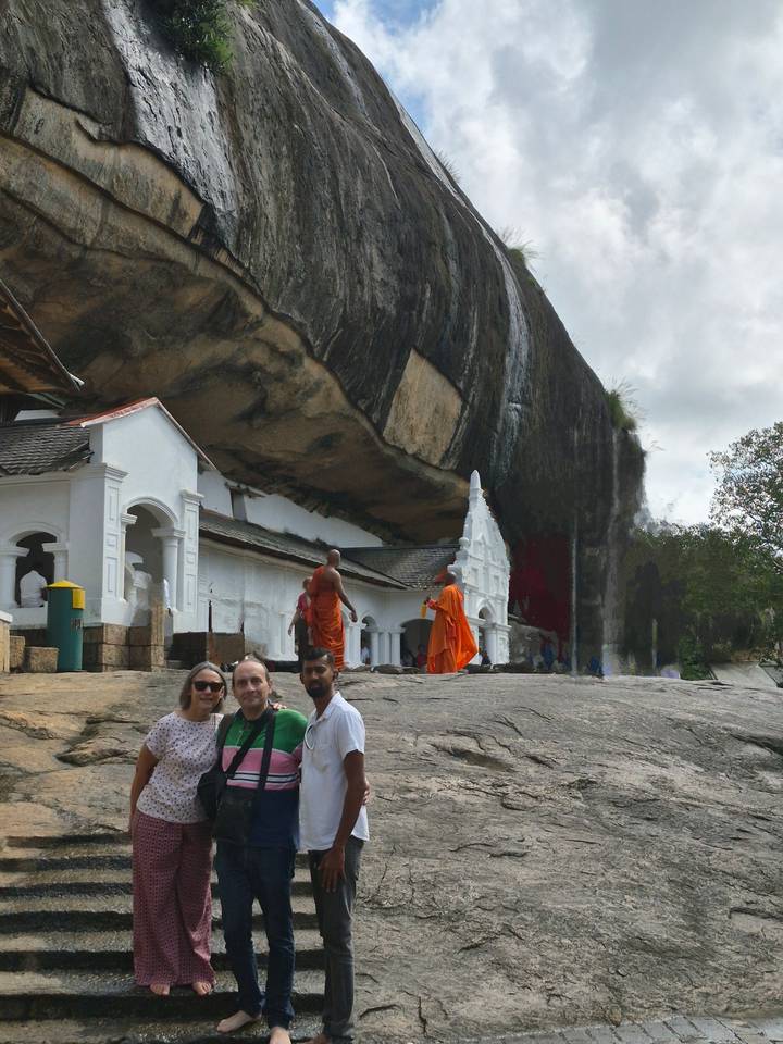 Travellers and robed monks at the entrance of a cave temple built beneath an overhanging rock.