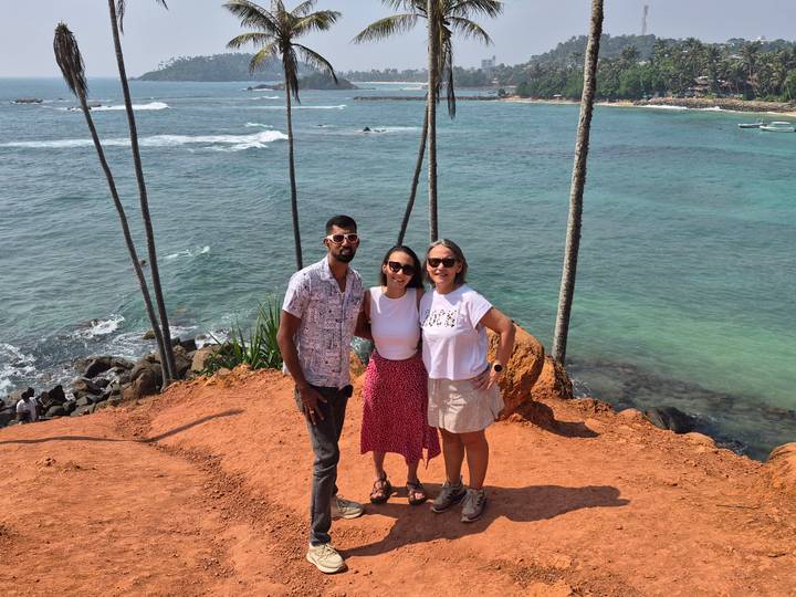 Three friends stand on red soil cliff with tall palm trees overlooking turquoise ocean waves.