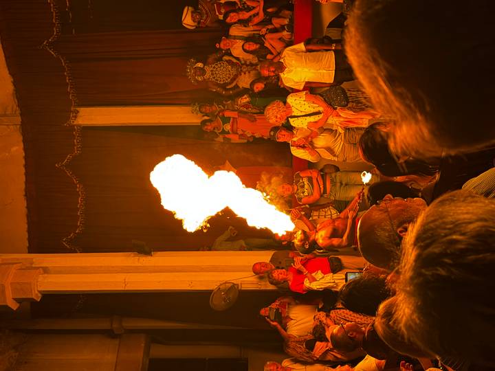 Performer breathes a plume of fire on stage while an audience watches inside a traditional hall.