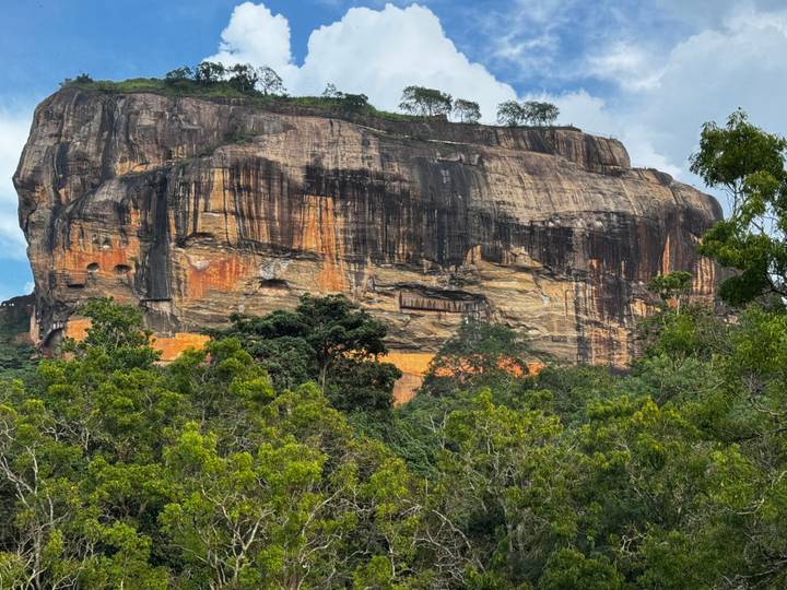 Massive Sigiriya rock fortress rises above dense jungle under a partly cloudy sky.