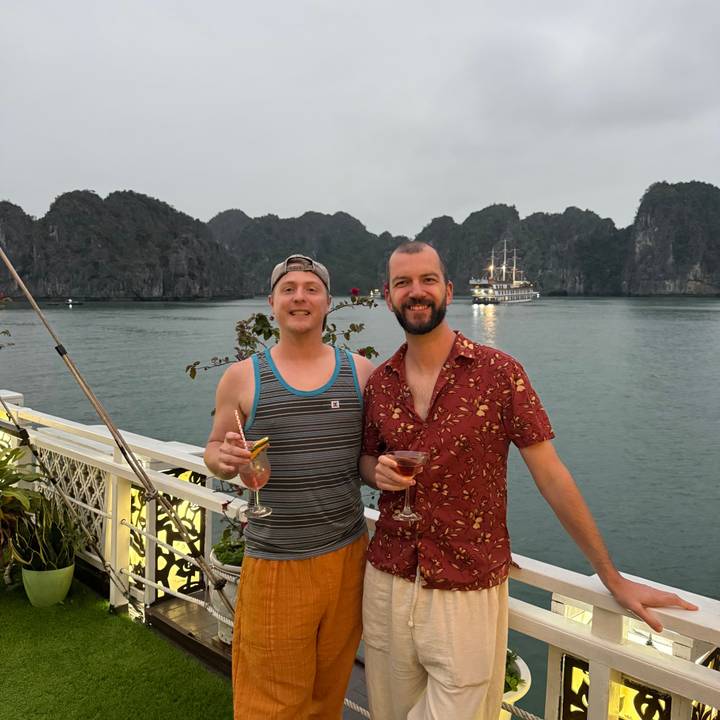 Two friends toast cocktails on a deck with limestone karst islands and a cruise ship in the background.